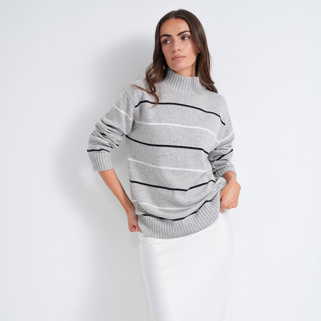 A woman wearing a light gray mock-neck knit sweater with horizontal black and white stripes, paired with a white skirt, standing against a light gray studio background.