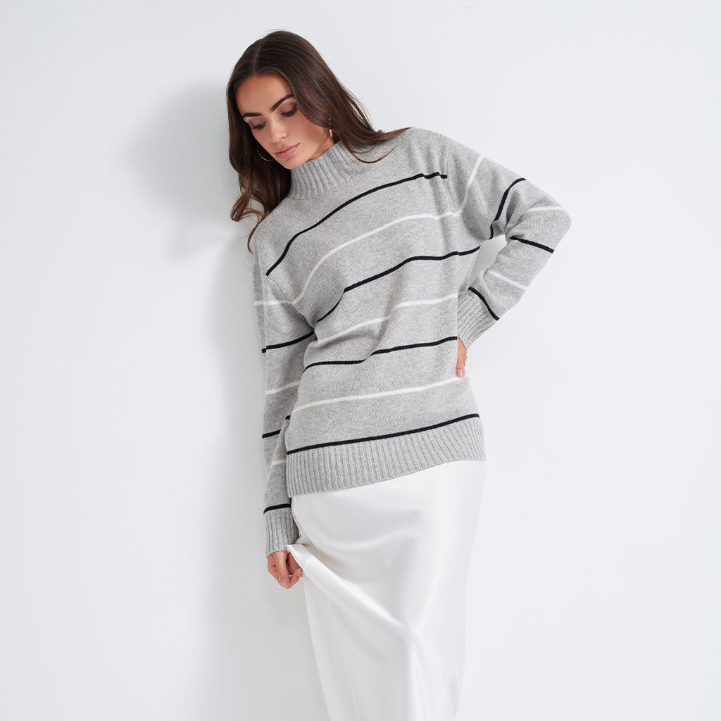 A woman wearing a light gray knit sweater with thin black and white horizontal stripes, paired with a white skirt, posing against a plain white studio background.