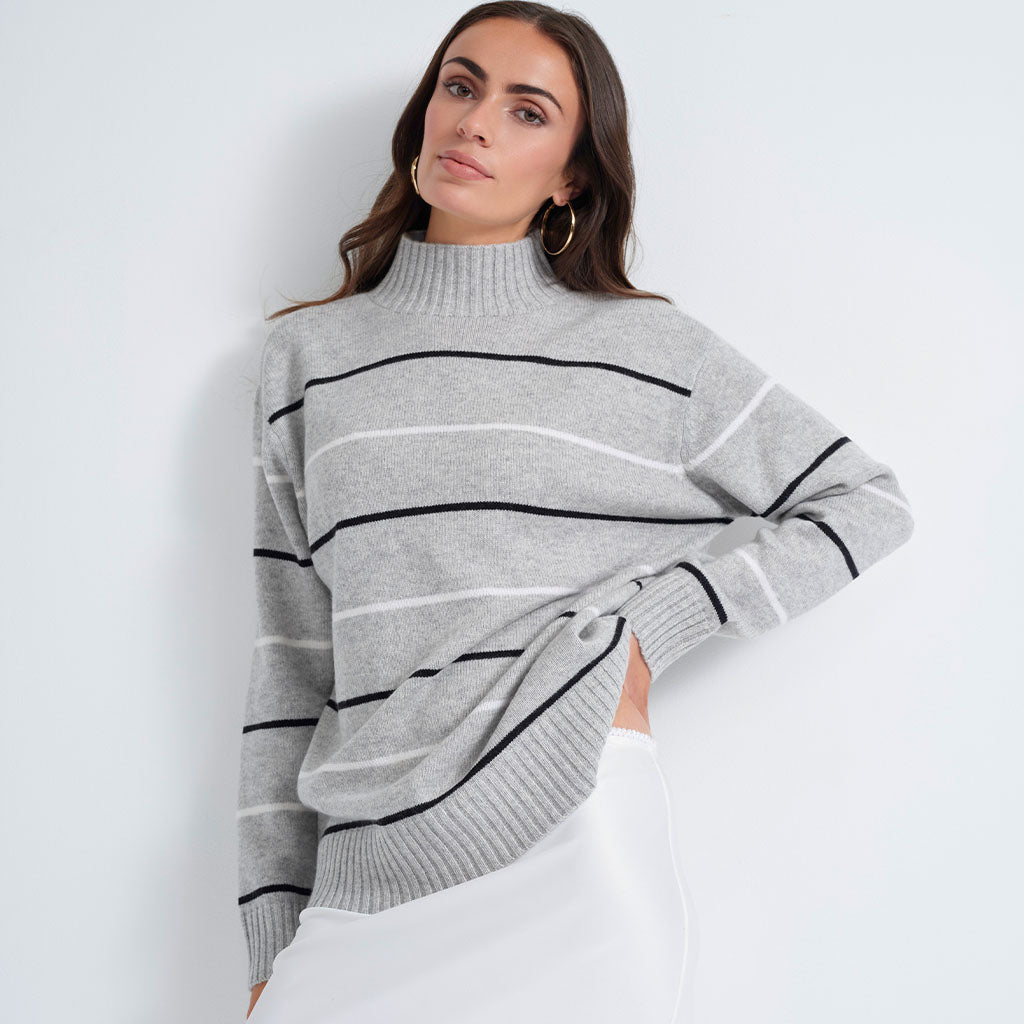 A woman wearing a light gray knit mock-neck sweater with horizontal black and white stripes, paired with white pants, posing against a plain white wall.