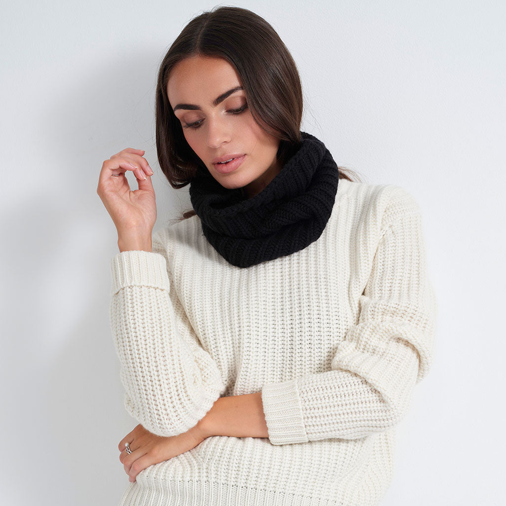 A woman wearing a thick black knit cowl neck warmer and a cream-colored ribbed knit sweater, posing against a light gray studio background.
