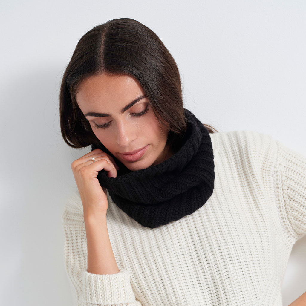 A woman wearing a thick black knit cowl neck warmer and a cream-colored textured knit sweater, posing against a light gray background.