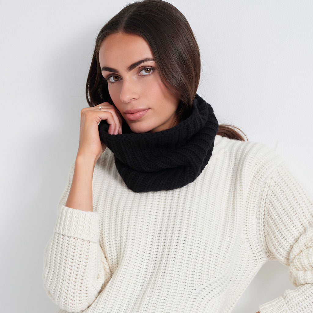 A woman wearing a cream knit sweater holds up a black cable-knit cowl neck warmer against a plain light gray background.