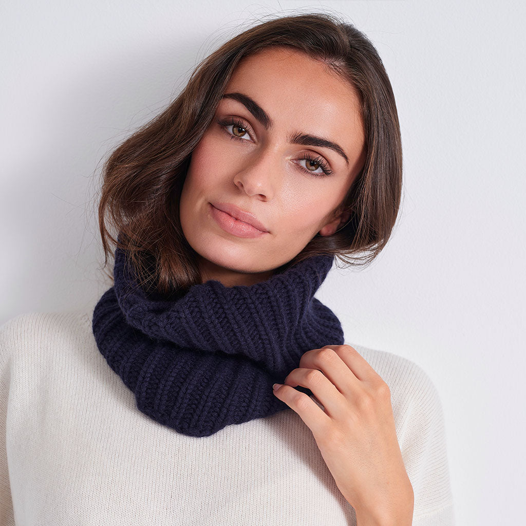 A woman wearing a thick, ribbed knit navy blue cowl neck warmer over a cream-colored sweater, photographed against a clean, light gray background.