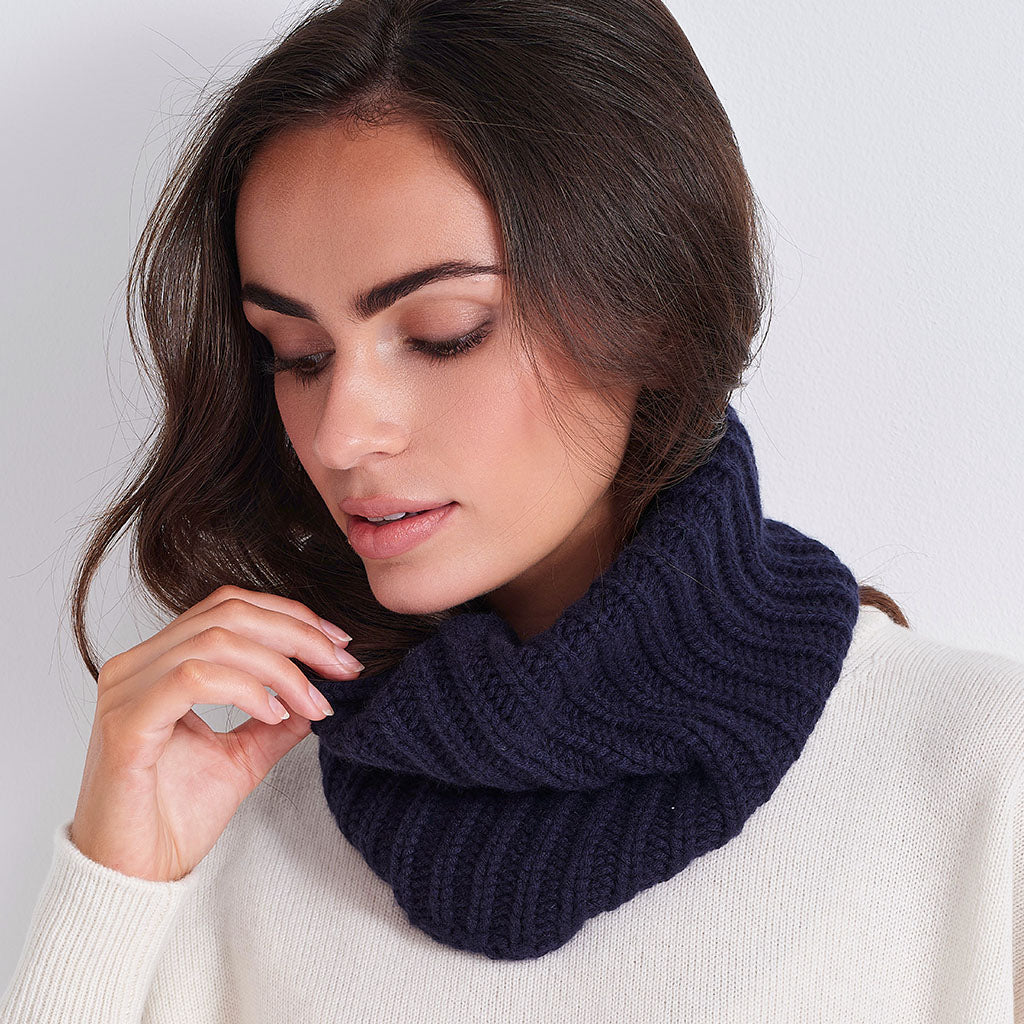 A woman wearing a dark navy blue ribbed knit cowl neck warmer over a cream-colored sweater, shown in a close-up portrait against a light gray background.