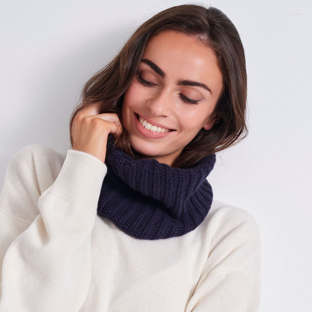 A woman wearing a dark navy blue ribbed knit cowl neck warmer over a cream-colored sweater, posing against a plain white background.