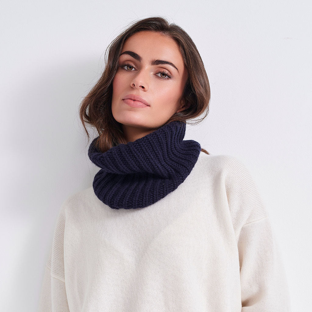 A woman wearing a thick, ribbed navy blue cowl neck warmer over a cream-colored knit sweater, photographed against a clean white background.