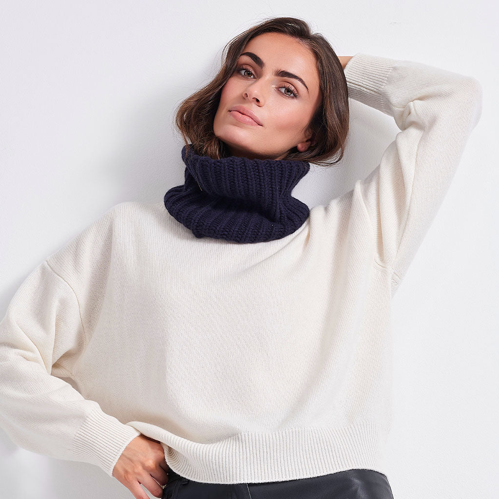 A woman wearing a thick, ribbed navy blue cowl neck warmer over a cream-colored knit sweater, photographed against a clean white background.
