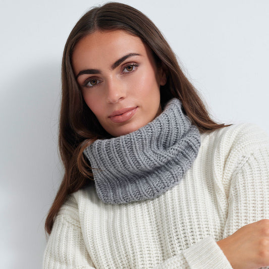A woman wearing a chunky gray ribbed knit cowl neck warmer over a cream-colored ribbed knit sweater, photographed against a light gray studio background.