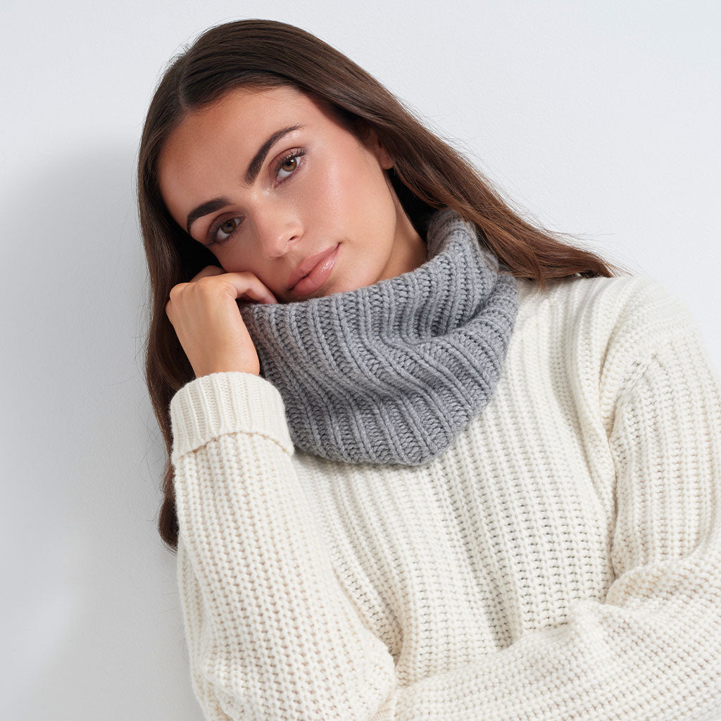 A woman wearing a gray ribbed knit cowl neck warmer over a cream-colored textured knit sweater, posing against a light gray background.