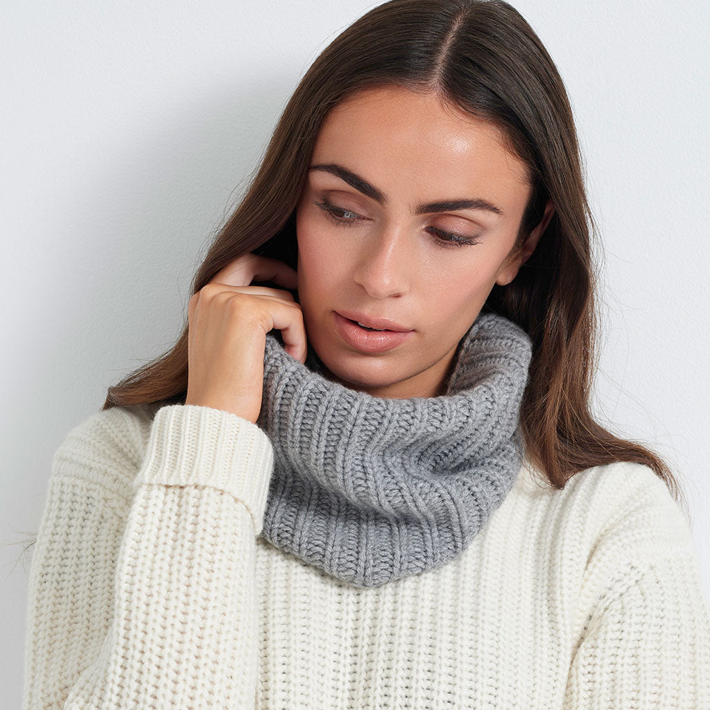 A woman wearing a gray ribbed knit cowl neck warmer over a cream-colored chunky knit sweater, photographed against a plain light gray background.