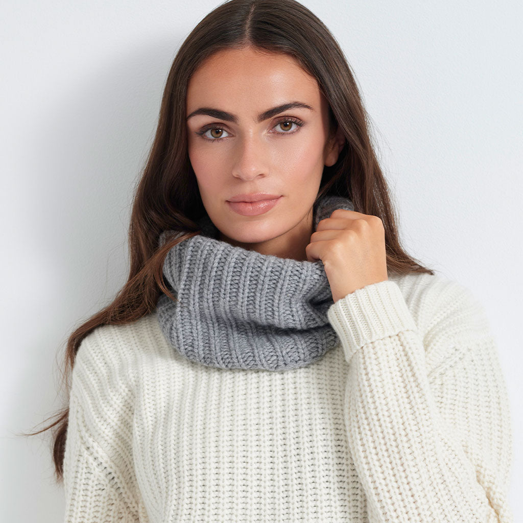 A woman wearing a chunky gray ribbed knit cowl neck warmer over an off-white ribbed knit sweater, photographed against a light gray studio background.