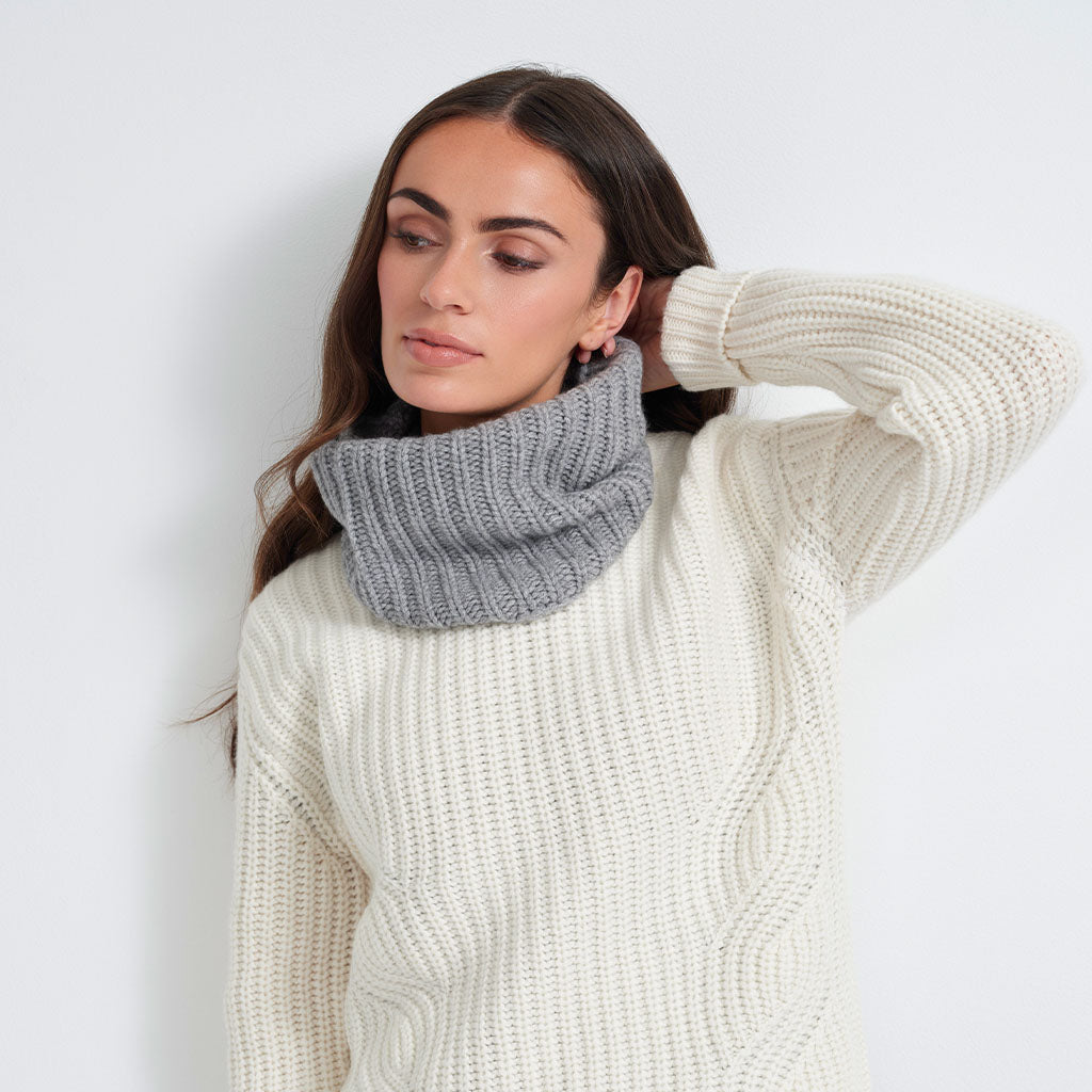 A woman wearing a gray ribbed knit cowl neck warmer over a cream cable-knit sweater, posing against a plain white wall.