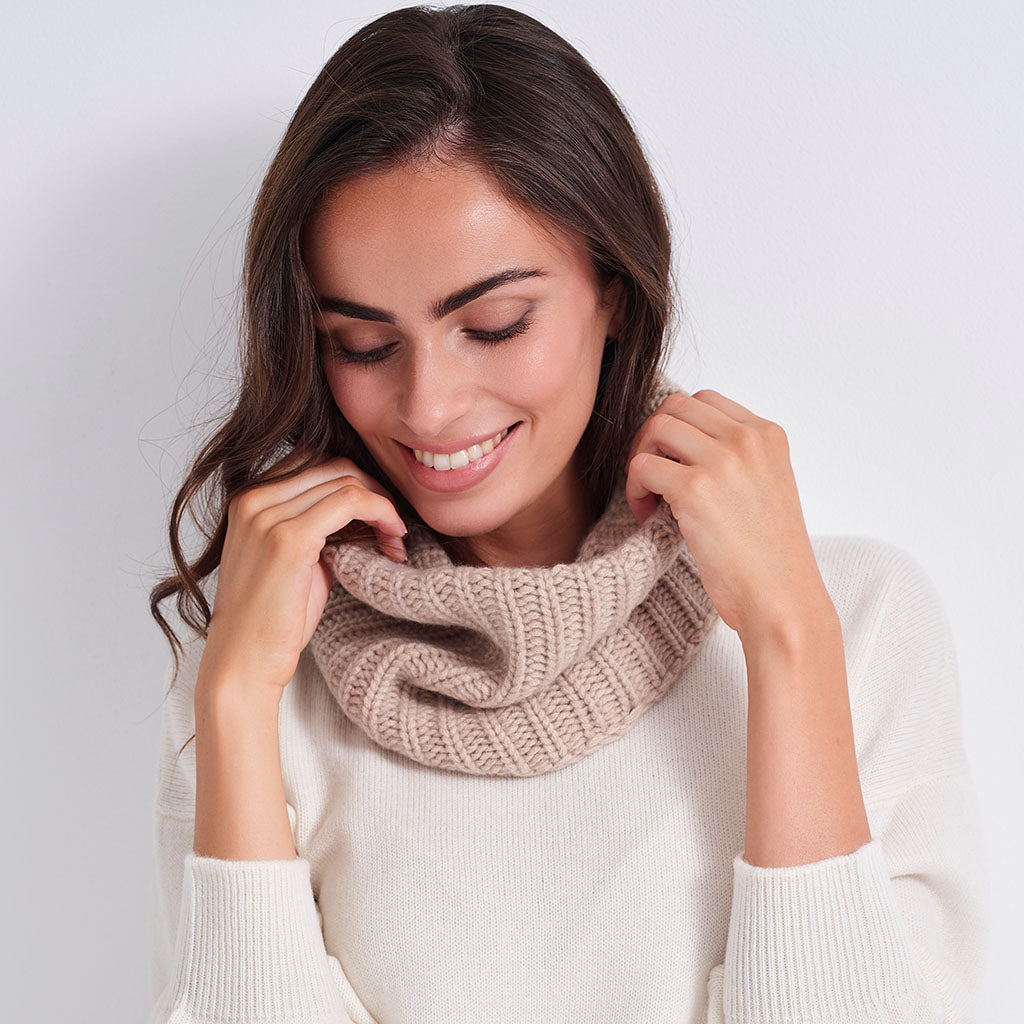 A woman wearing a cream-colored knit sweater and a beige ribbed cowl scarf, posing against a clean white studio background.