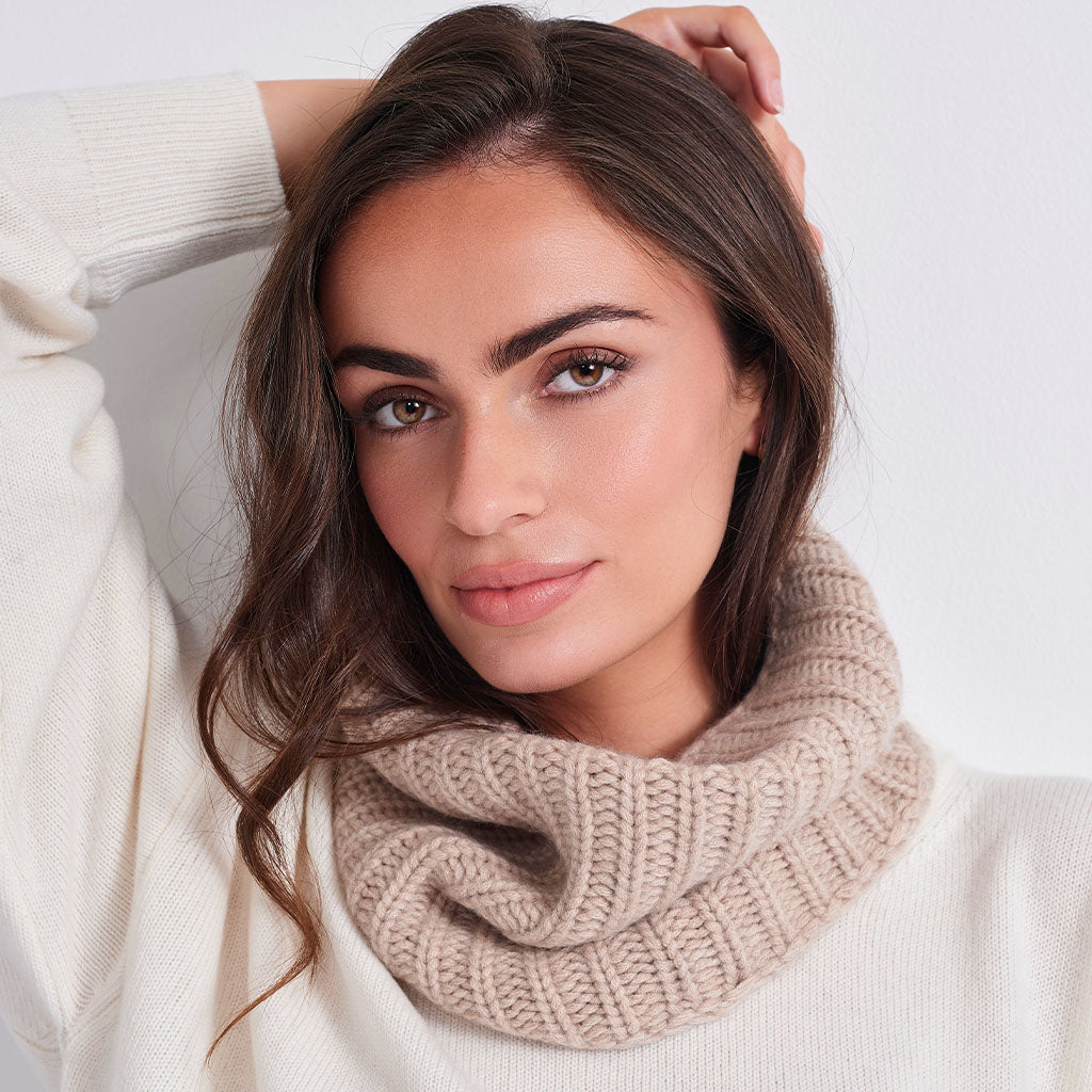 A woman with long brown hair wearing a cream-colored knit sweater and a beige ribbed cowl neck warmer, posing against a clean white background.