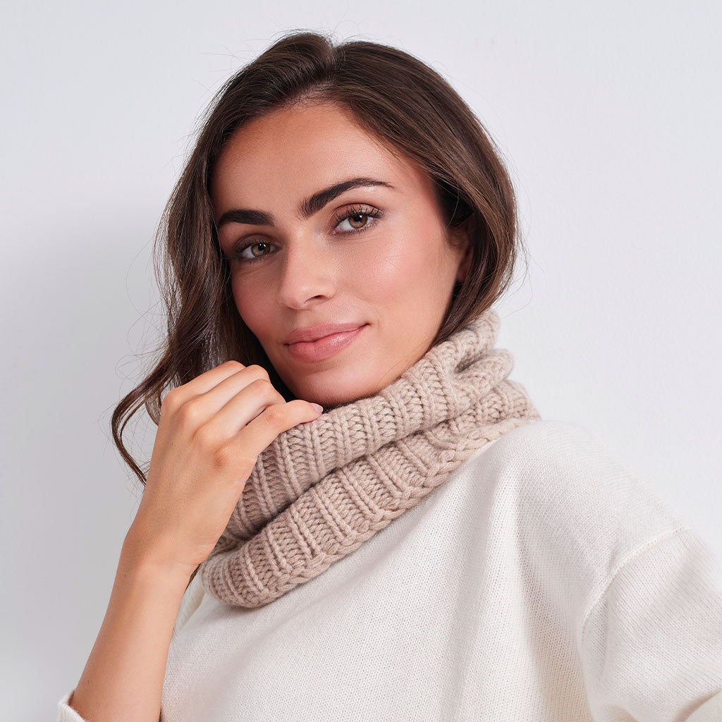 A woman wearing a chunky ribbed knit cowl scarf in a light beige color, paired with a cream-colored knit sweater, against a plain light gray background.