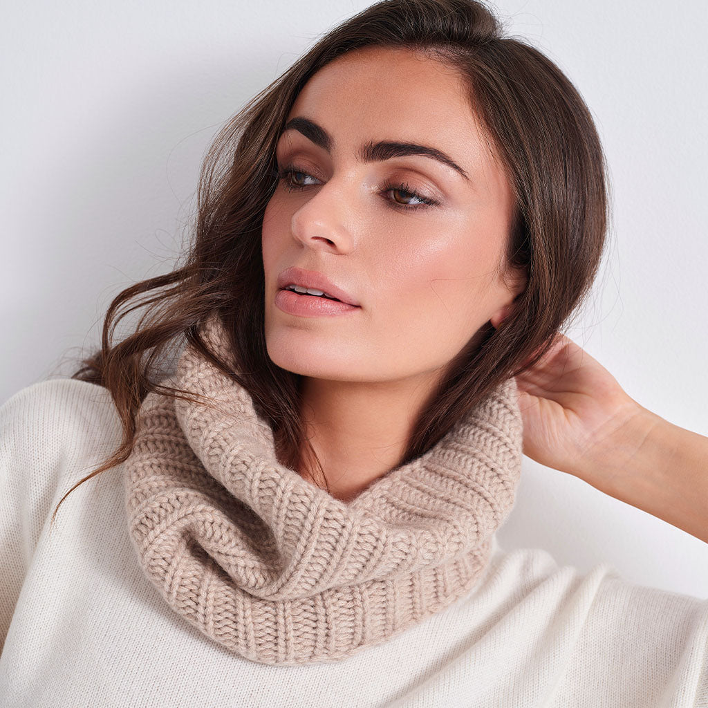 A woman wearing a chunky knit beige cowl neck warmer over a cream-colored sweater, shown in a close-up portrait against a plain light background.