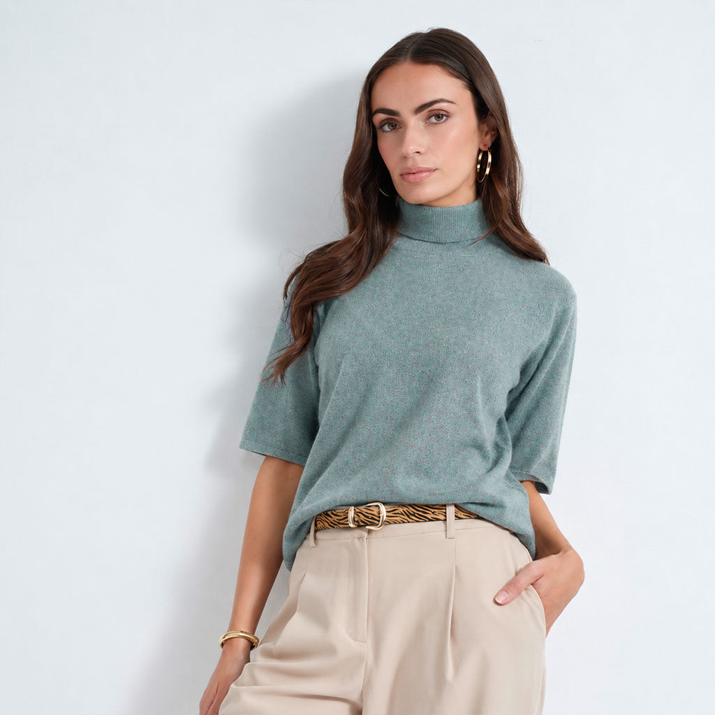 A woman modeling a lagoon short-sleeve mock neck knit top with light beige trousers and a patterned belt, standing against a light blue-gray studio background.