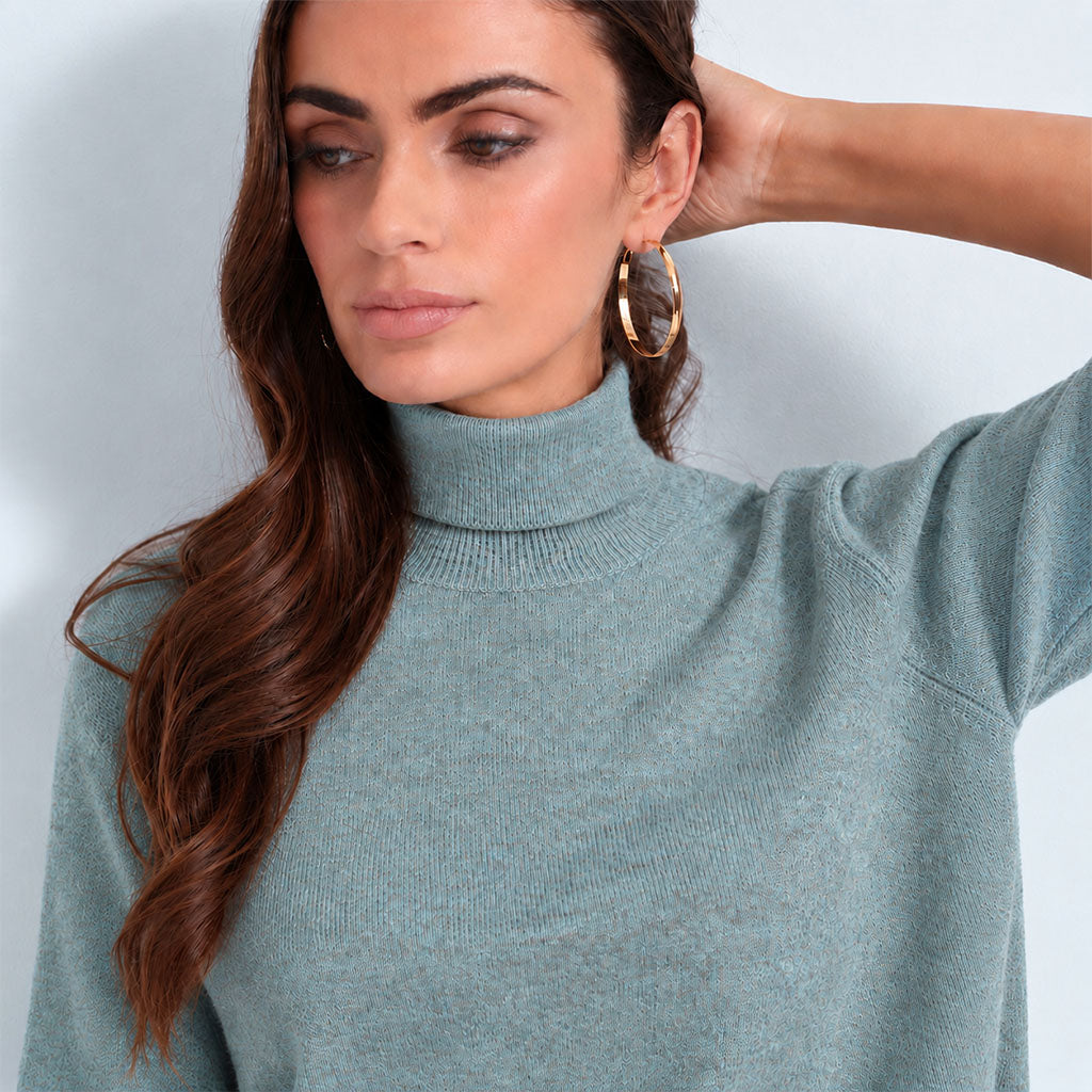 A woman wearing a light blue-gray ribbed turtleneck sweater and large gold hoop earrings, shown in a close-up studio shot against a pale gray background.