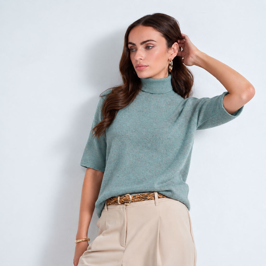 A woman modeling a lagoon short-sleeve mock neck knit top with light beige trousers and a patterned belt, standing against a light blue-gray studio background.