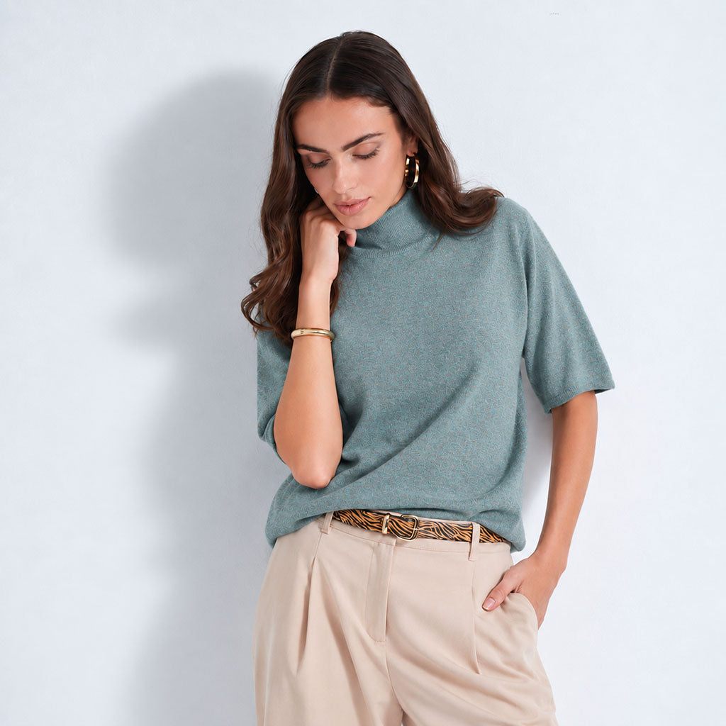 A woman modeling a lagoon short-sleeve mock neck knit top with light beige trousers and a patterned belt, standing against a light blue-gray studio background.