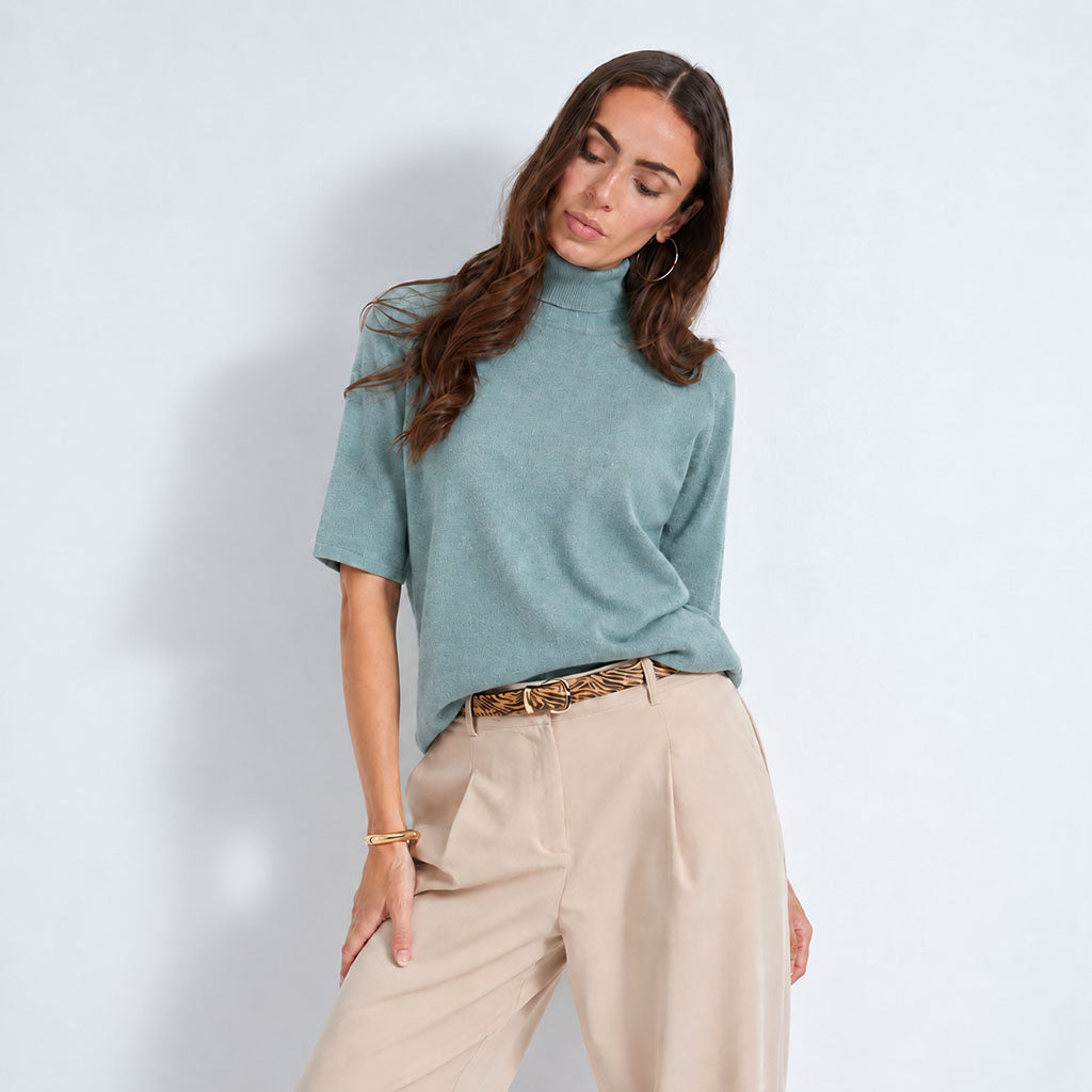A woman modeling a lagoon short-sleeve mock neck knit top with light beige trousers and a patterned belt, standing against a light blue-gray studio background.
