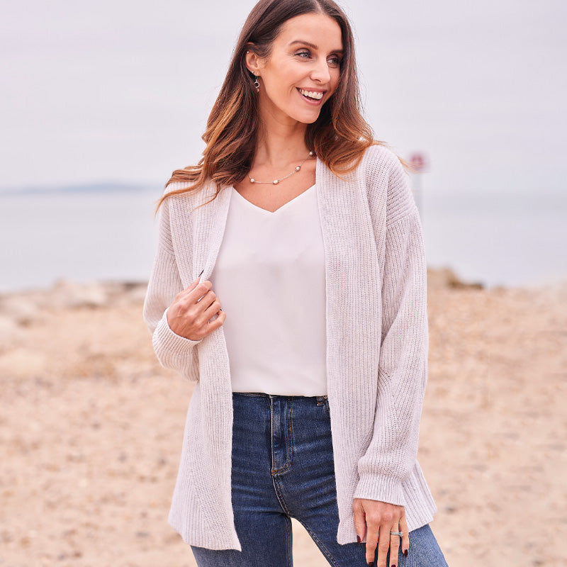 Woman wearing a cashmere light gray cardigan over a white shirt and blue jeans on a beach.