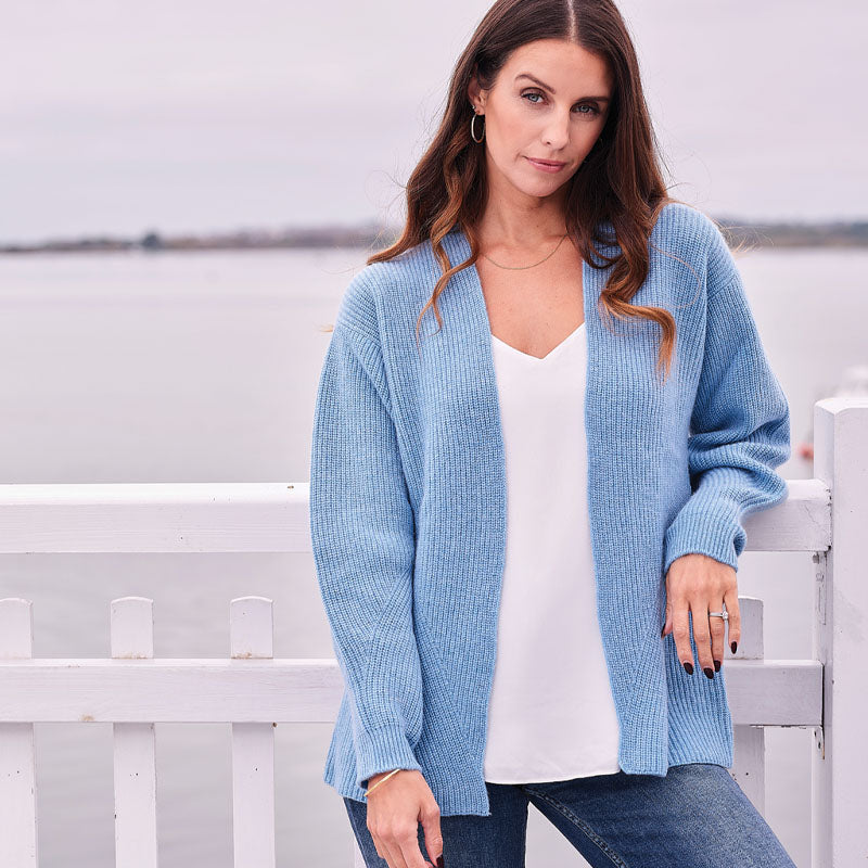 Woman wearing a cashmere light blue cardigan over a white top, standing in front of a white picket fence.
